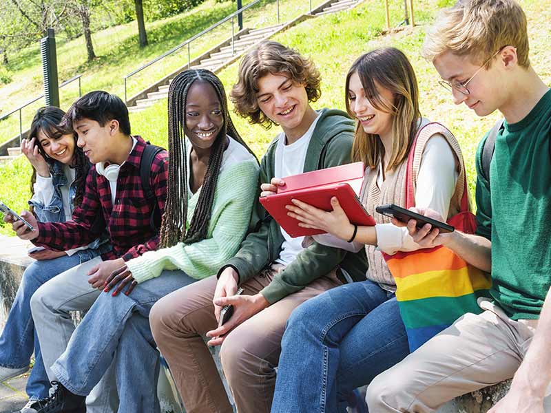 Multicultural Teenagers Interacting at School Park - Diverse teens sit on a wall, conversing and using mobile devices